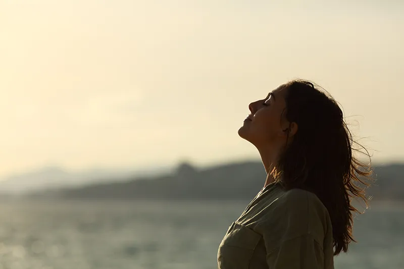 Eine ruhige Frau steht bei Sonnenuntergang am Meer, die Augen geschlossen, den Kopf leicht nach hinten geneigt, und genießt die Brise. Es wird ein Moment der Gelassenheit und Ruhe vermittelt.