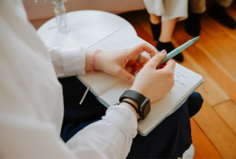 Nahaufnahme einer Person, die einen Stift hält und in ein Notizbuch auf ihrem Schoß schreibt. Eine weitere Person ist im Hintergrund verschwommen zu sehen. Die Szene wirkt ruhig und konzentriert.